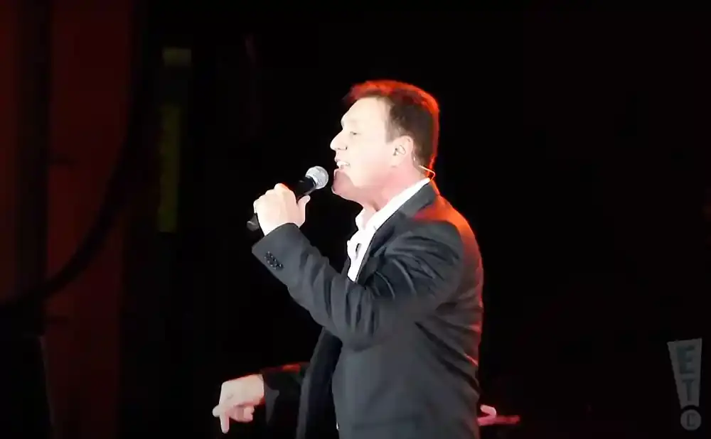 A man in a suit sings into a microphone on stage with red and black lighting.
