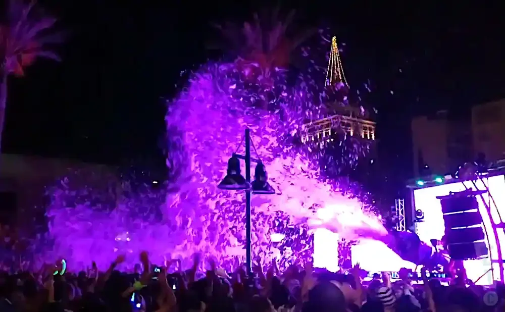 A crowd at a nighttime event watches a large plume of purple foam shoot from a stage.