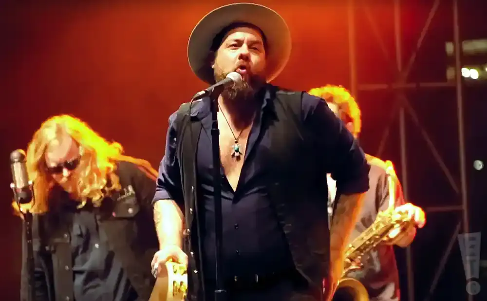 A man in a hat sings into a microphone on stage with a band.