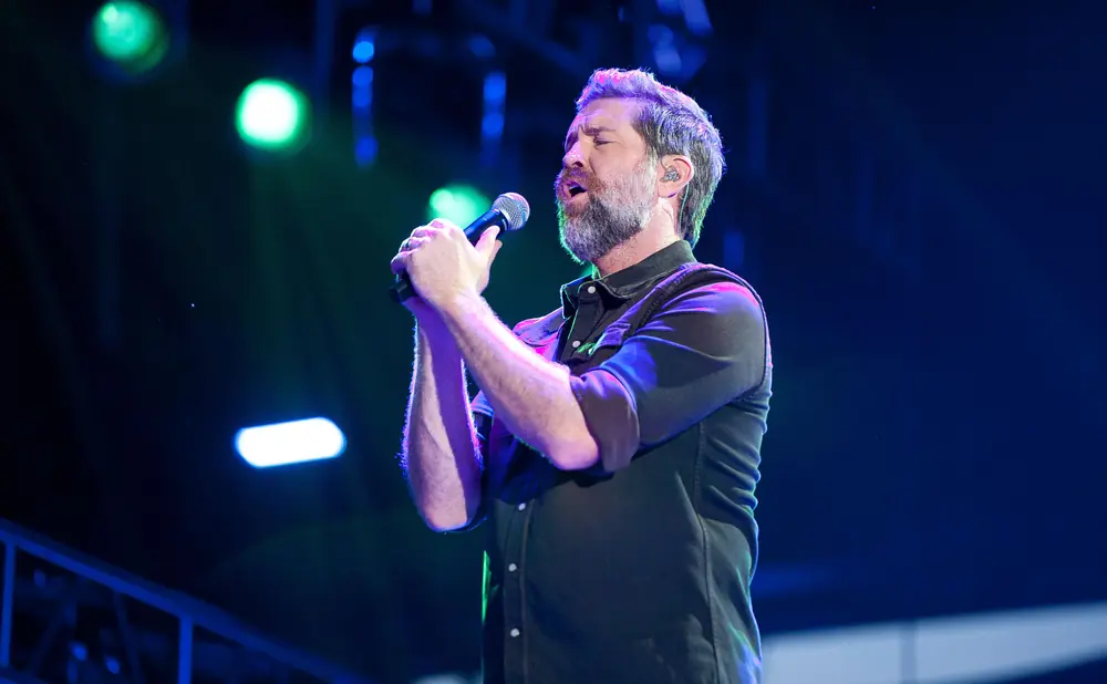 A man with a beard sings into a microphone on a stage with blue and green lights.