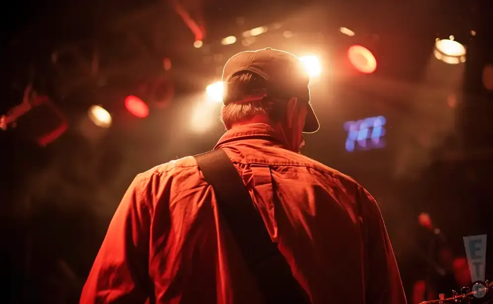 Back of a person wearing a baseball cap and orange shirt on stage with bright lights.