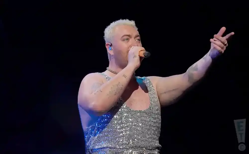 Singer Sam Smith performs on stage, wearing a sparkly silver tank top and holding a microphone.