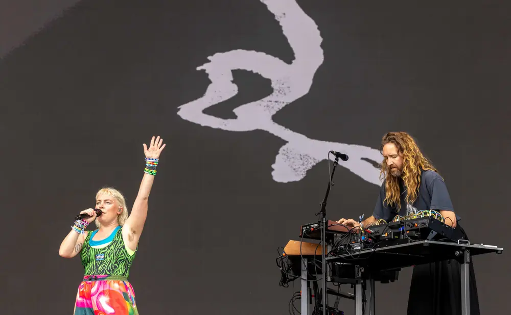 Singer with colorful outfit and bracelets sings into microphone, while a man with long blond hair plays electronic equipment.