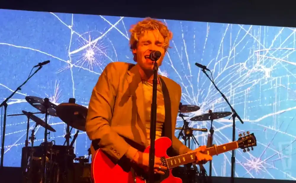 A male singer with blonde curly hair plays a red electric guitar and sings into a microphone on stage with a blue cracked screen background.