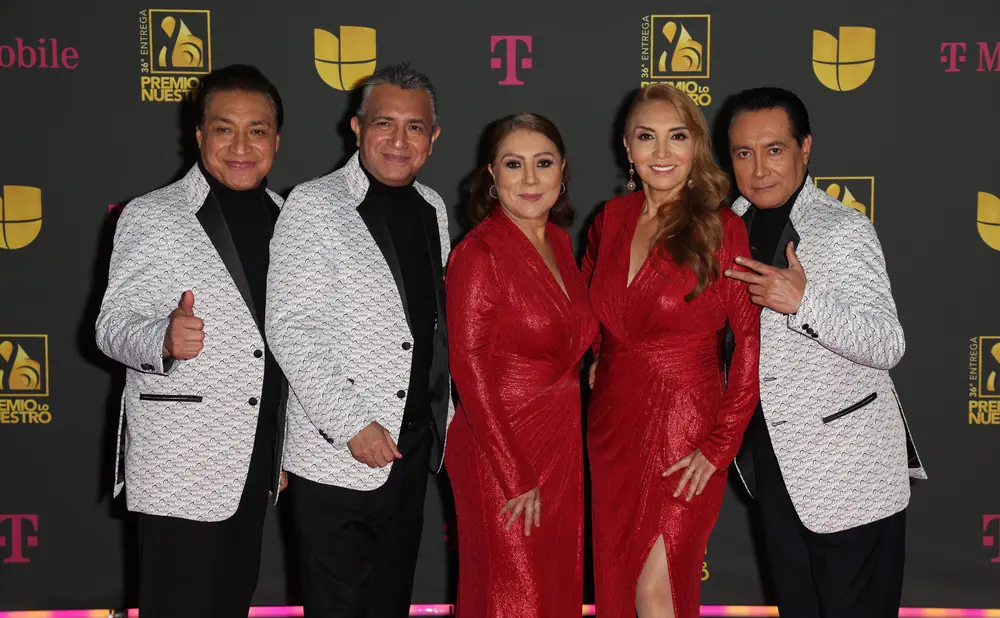 A group of four people, two women in red dresses and two men in patterned jackets, pose on a pink carpet.