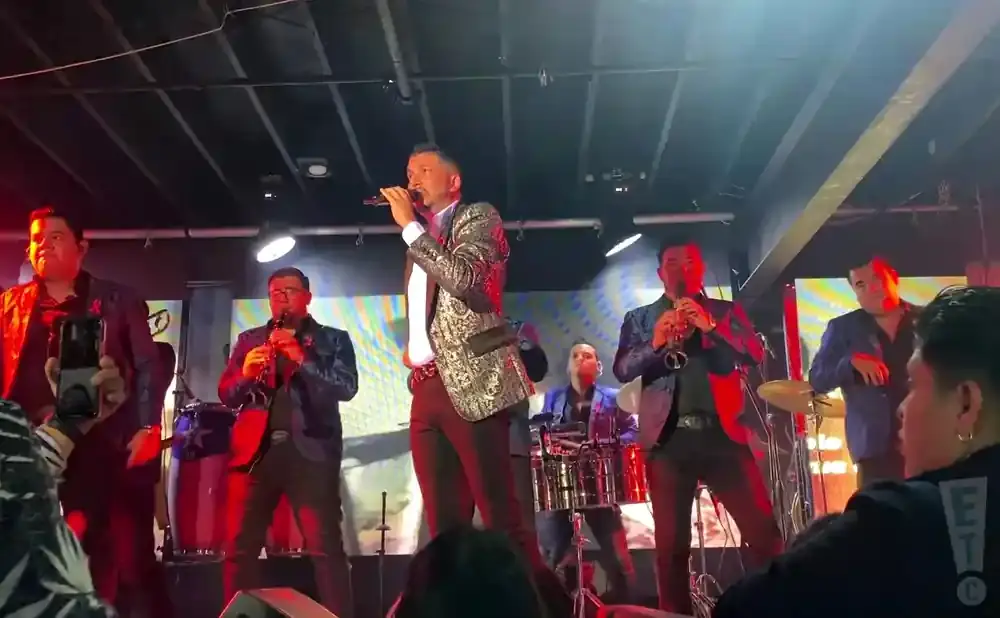 A male singer in a sequined jacket performs on stage with a band playing brass instruments and drums.