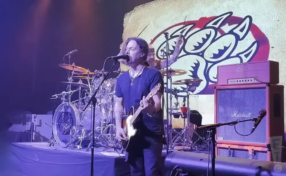 A man plays guitar and sings on stage with a drum set and Marshall amps behind him.