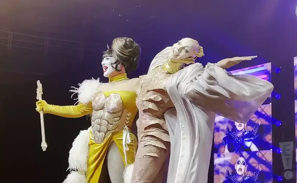 Two drag performers on stage, one in a sparkling yellow outfit with a feather boa and a staff, the other in a draped white costume.