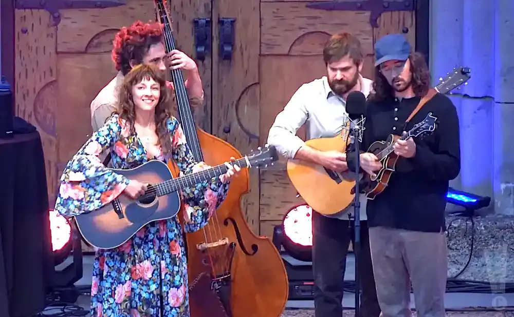 A band performs bluegrass music: a woman on guitar, a man on bass, and two men playing mandolins.
