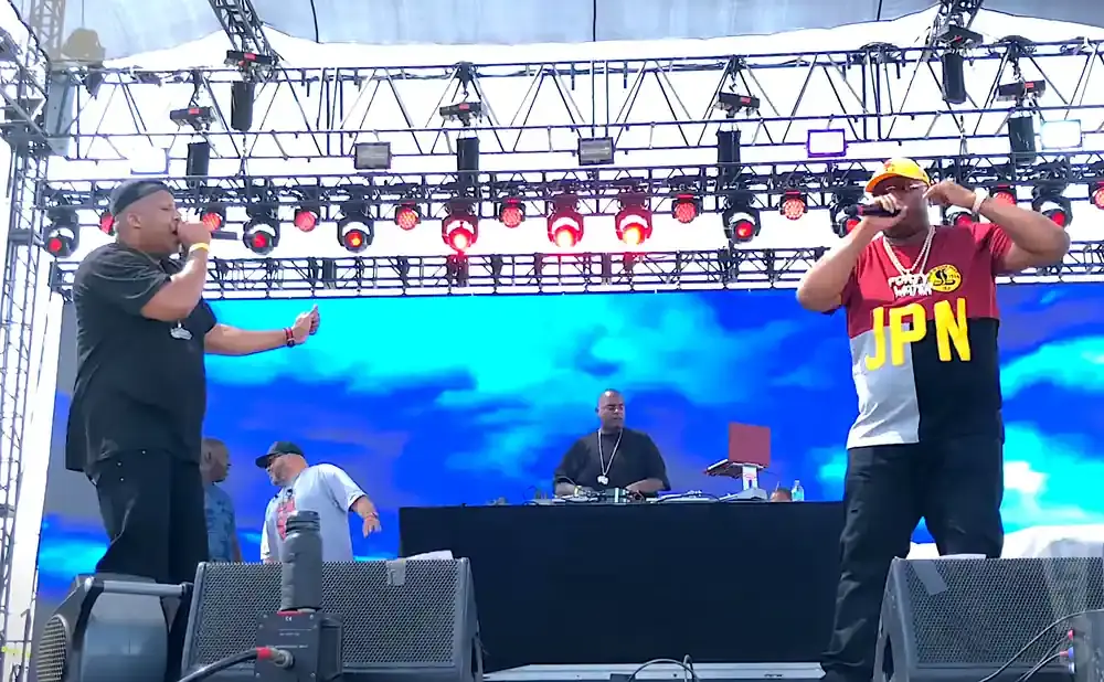 Rappers perform on stage with a DJ and a backdrop of blue clouds.