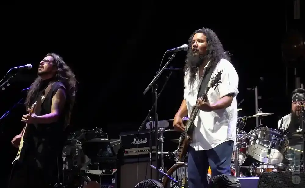 A band performs on stage with a dark background. Two men are visible playing guitars and singing into microphones.