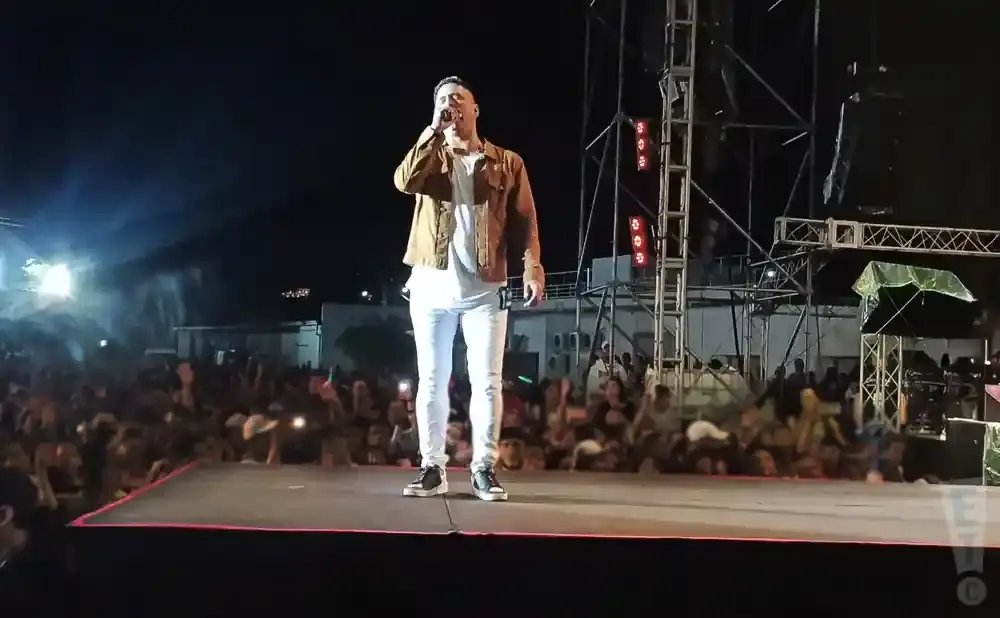 A male singer in a brown jacket and white pants performs on stage for a large crowd at night.