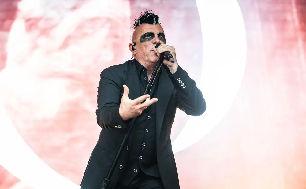 A man with a mohawk and dark eye makeup sings into a microphone on a stage, wearing a black suit.