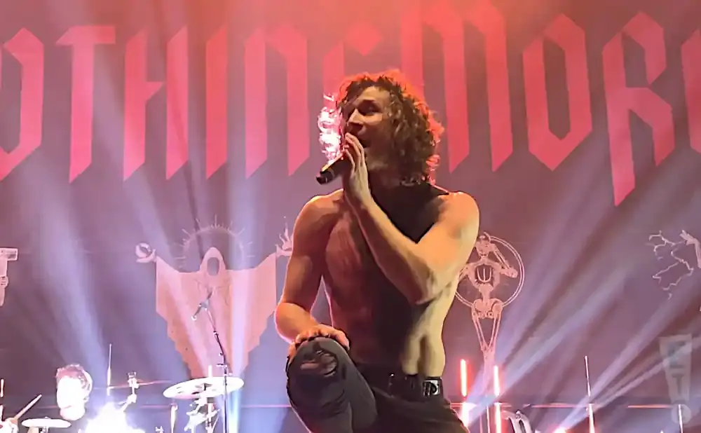 Shirtless male singer with curly hair performs on stage with a banner behind him.