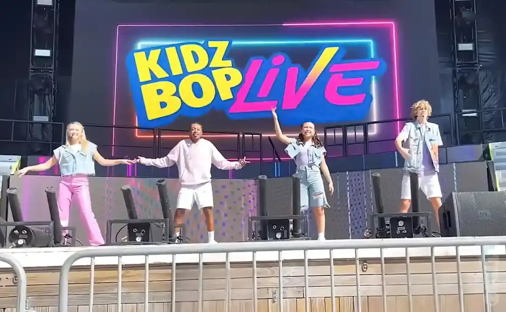 Four Kidz Bop Live performers dance on stage in front of a bright neon sign.