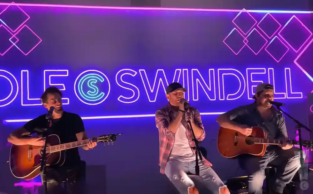 Cole Swindell performing with two other musicians on stage with a neon sign in the background.