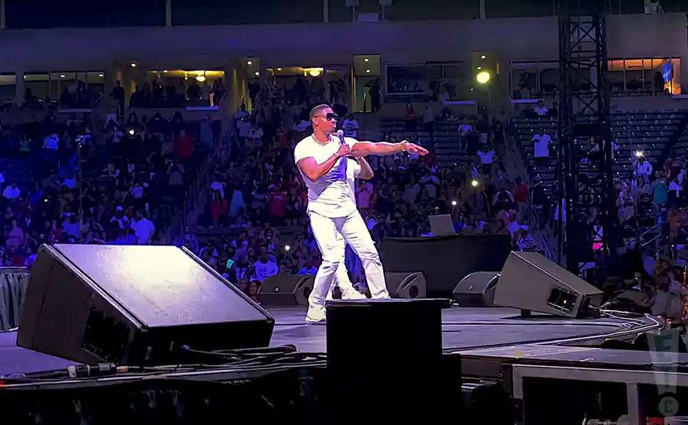 A male performer in white clothing sings into a microphone on a concert stage, facing a cheering crowd in stadium seating.