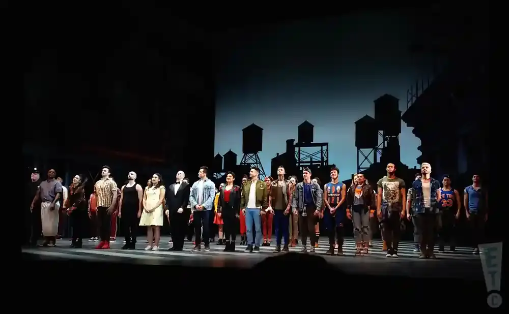 A diverse cast of performers stands on stage, posing for a photo in front of a New York City skyline backdrop.