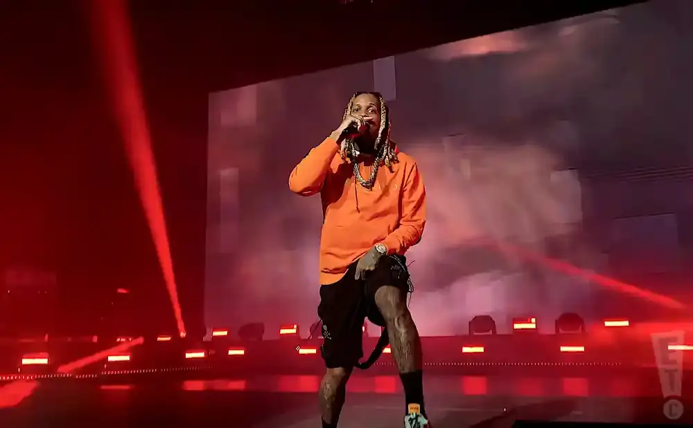 Rapper Lil Durk performs on stage in an orange hoodie and black shorts, illuminated by red lights.