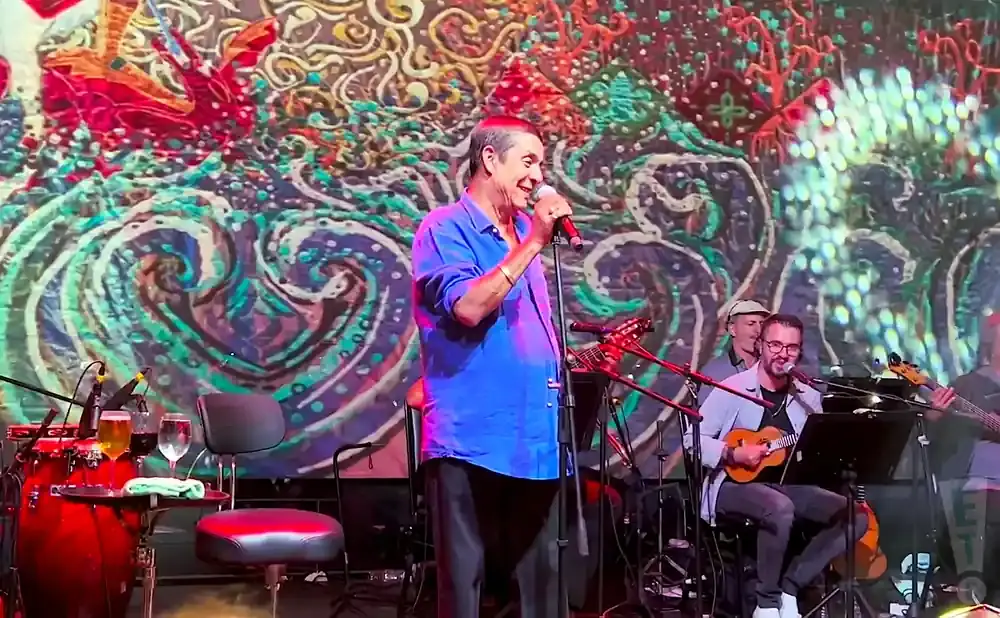 Singer in a blue shirt performs with a band on a stage with a colorful, abstract backdrop.