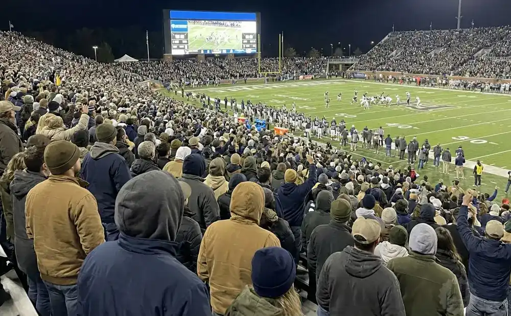 Spectators watch a night football game from stadium bleachers, with a large scoreboard visible.