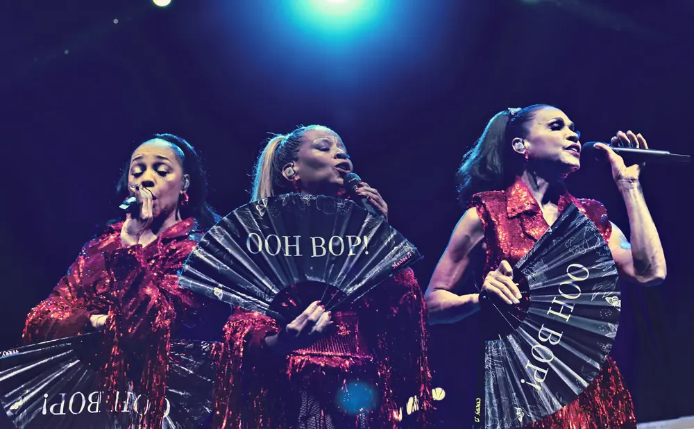 Three women in sparkly red outfits sing into microphones, holding black fans with white text.