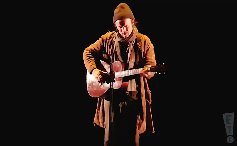 A man wearing a beanie and a long coat plays an acoustic guitar on a dark stage.