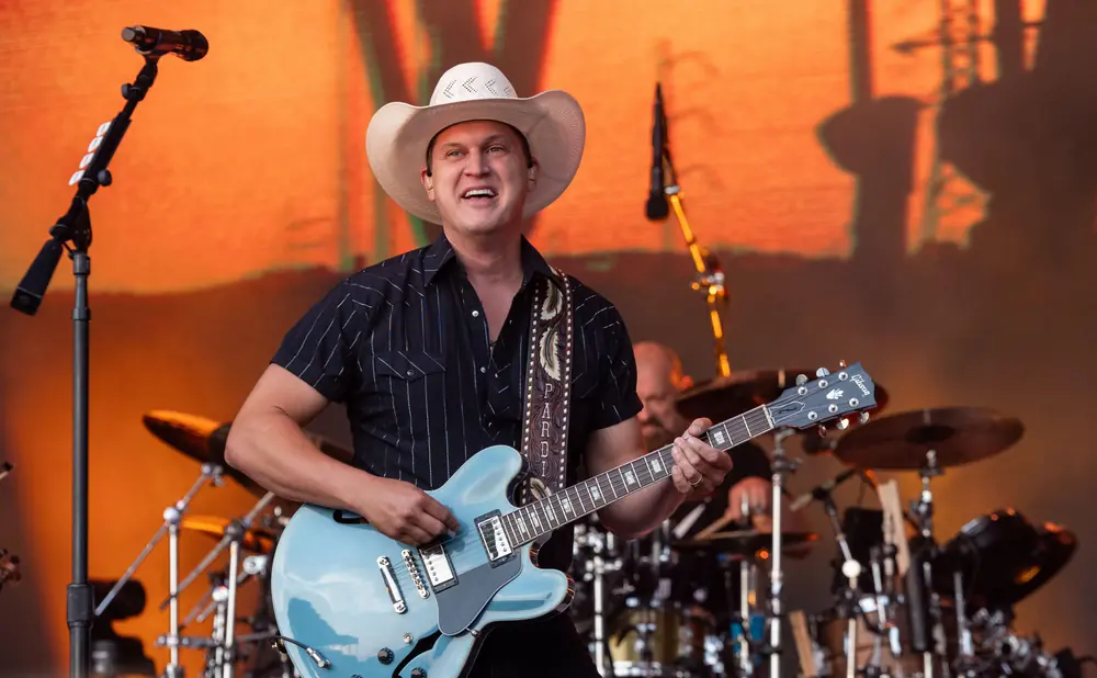 A man in a cowboy hat plays a light blue electric guitar on stage with a band.