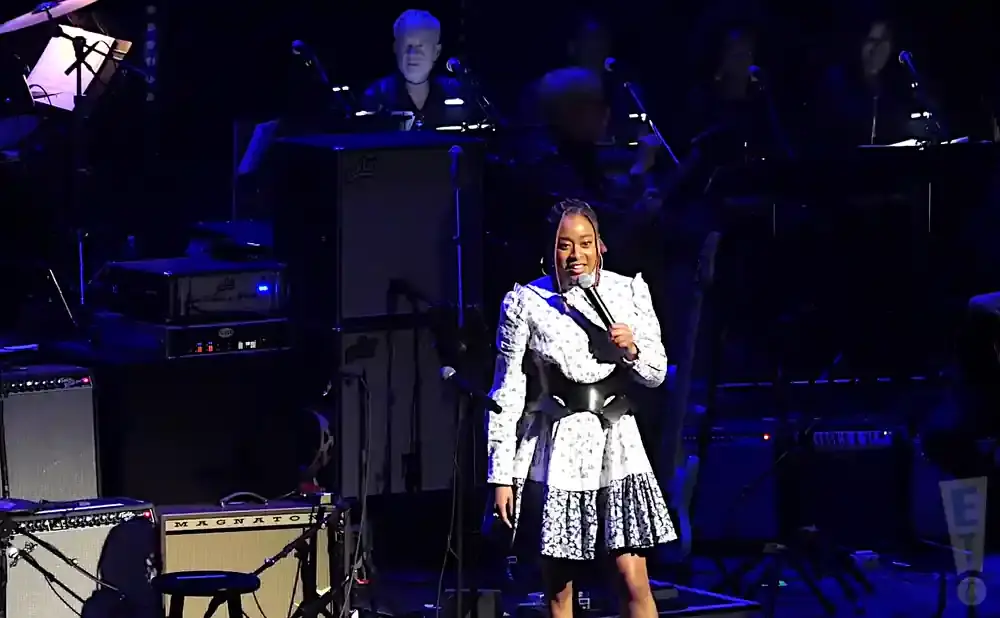 A Black woman in a floral dress sings into a microphone on a stage with musical equipment.