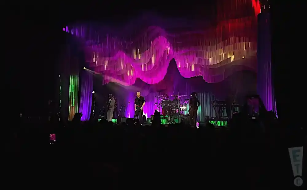 A band performs on stage with a wavy, colorful backdrop and dramatic lighting.