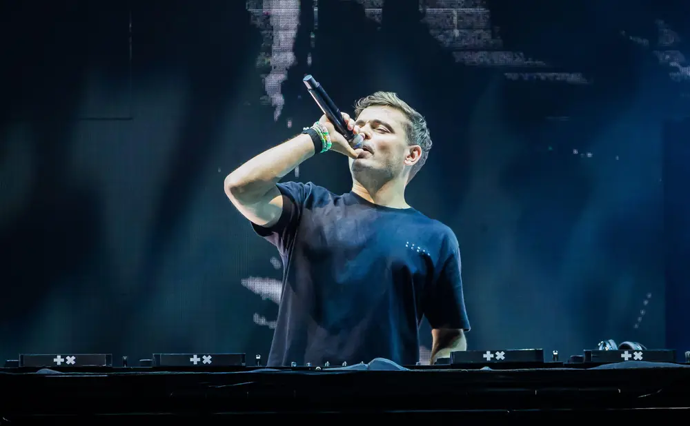 DJ Tiësto performing live on stage, singing into a microphone at a DJ booth with a large screen behind him.