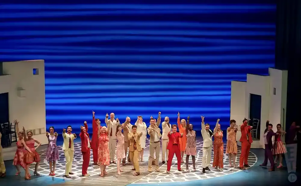 A cast of singers and dancers in colorful outfits strike a pose on a stage with a blue backdrop.