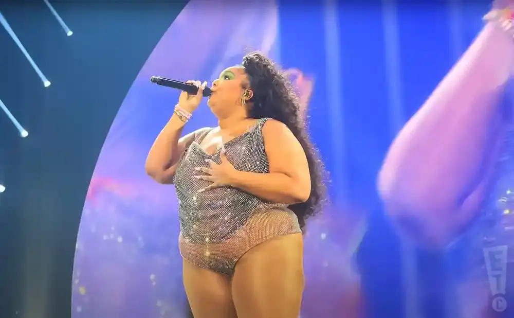 Singer Lizzo performs on stage in a sparkly leotard, holding a microphone and her hand on her chest.