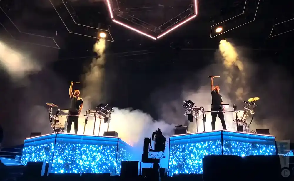 Two drummers on glowing blue stages, holding drumsticks aloft amidst stage smoke and lights.
