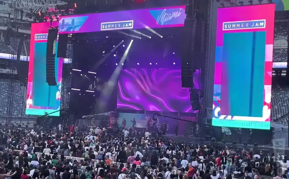 A large crowd at an outdoor music festival, with a big stage featuring screens and lights.