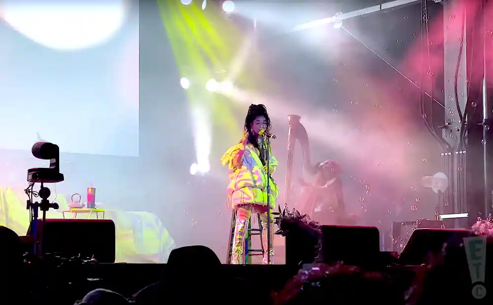 A woman in a puffy, colorful jacket sings into a microphone on a stage, with lights and bubbles in the background.