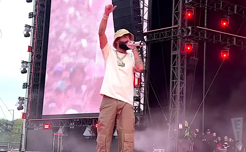 A male performer on stage, wearing a bucket hat and holding a microphone, with a large screen and red lights behind him.