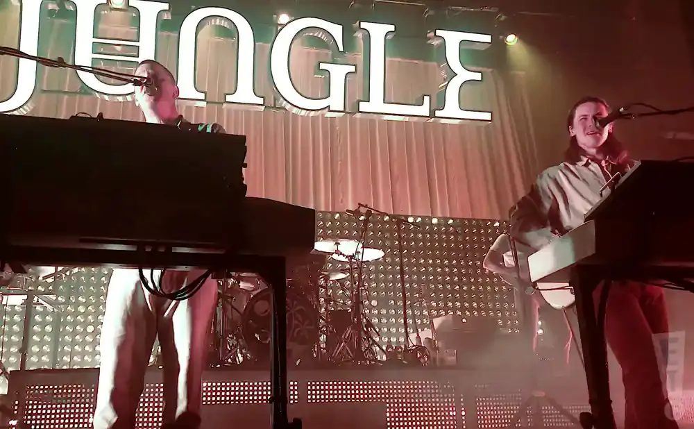 Two men playing keyboards on stage in front of a backdrop with the word 