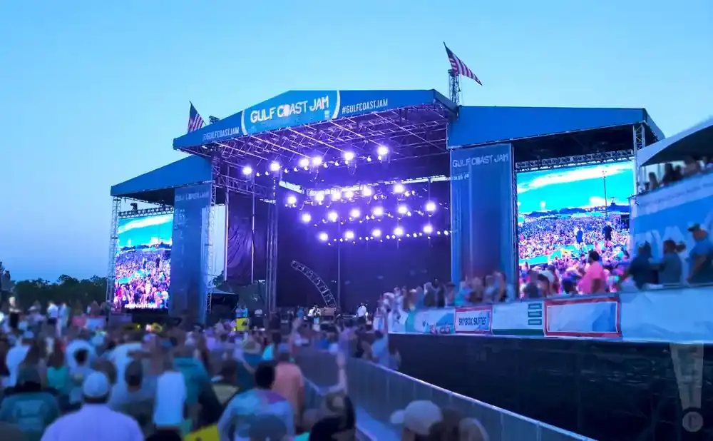 Outdoor concert stage with large screens displaying a crowd at 