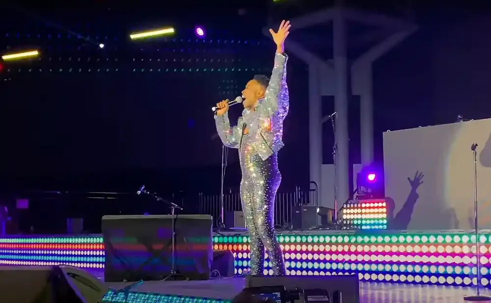 Performer in sparkly silver outfit singing into a microphone with arm raised on a brightly lit stage.