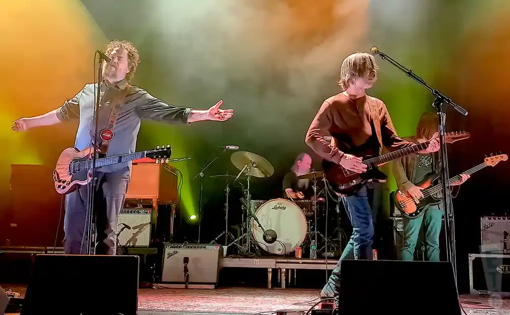 A band plays on stage, with a singer in the foreground holding a guitar and extending his arms.