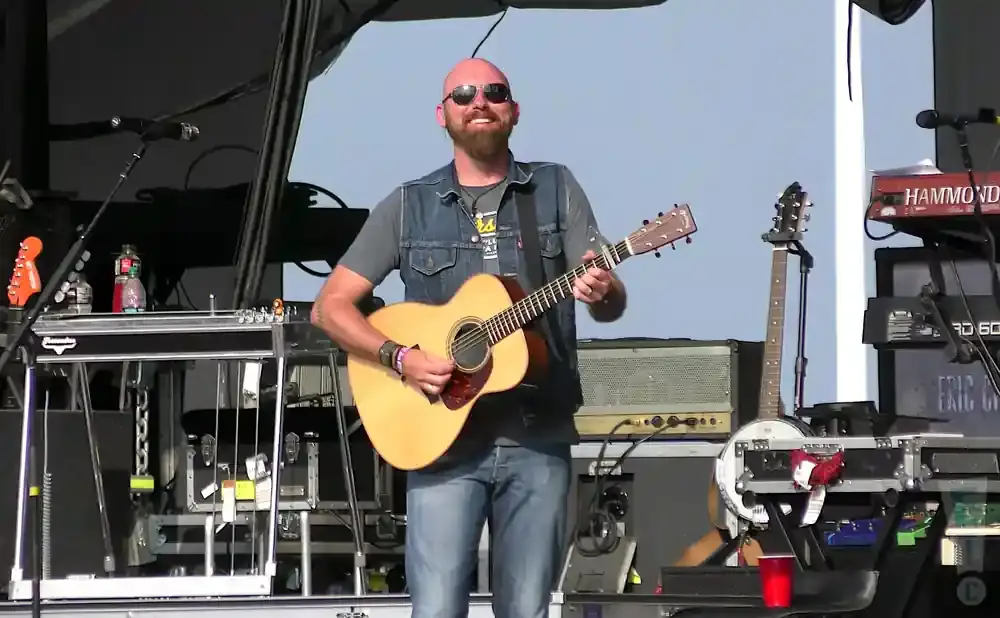 A bald man with a beard and sunglasses plays an acoustic guitar on stage.