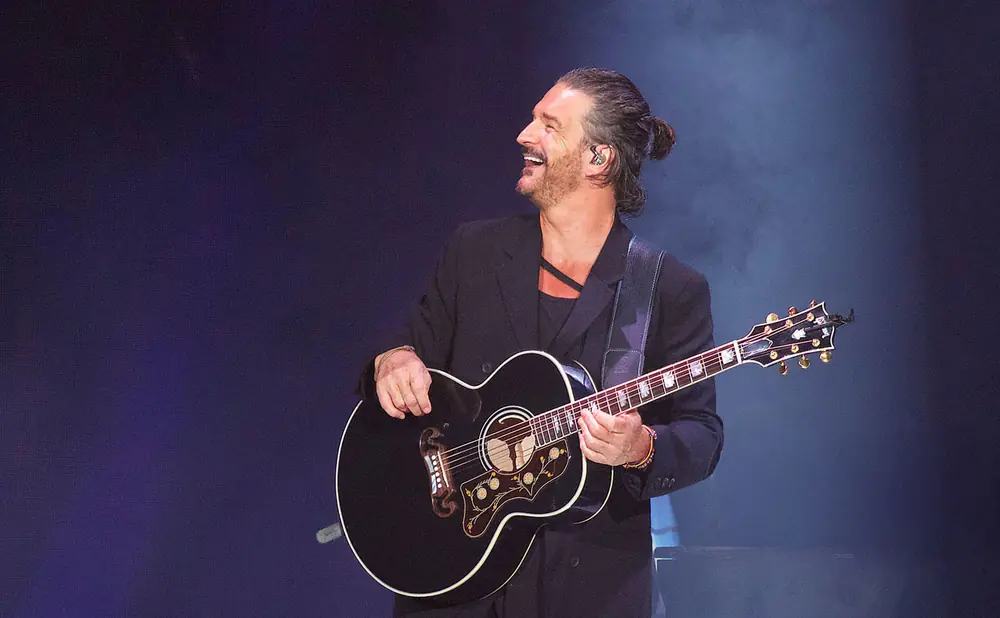 A smiling man with a bun in his hair plays an acoustic guitar on stage.