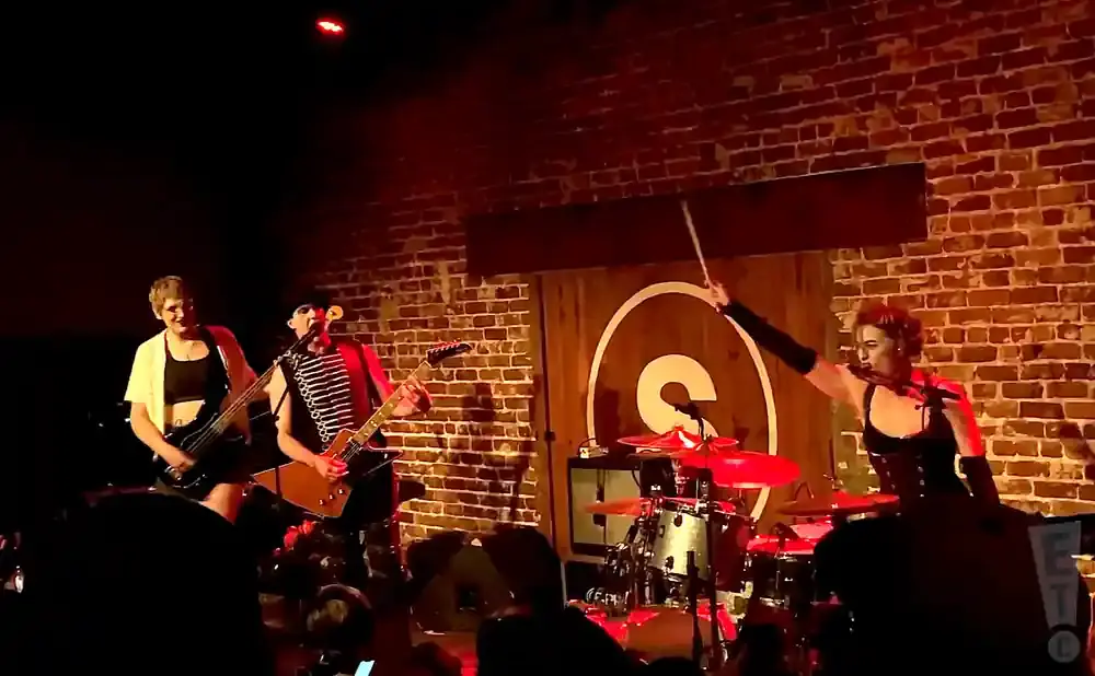 A band performs on a brick stage, with a bassist, guitarist, and drummer.
