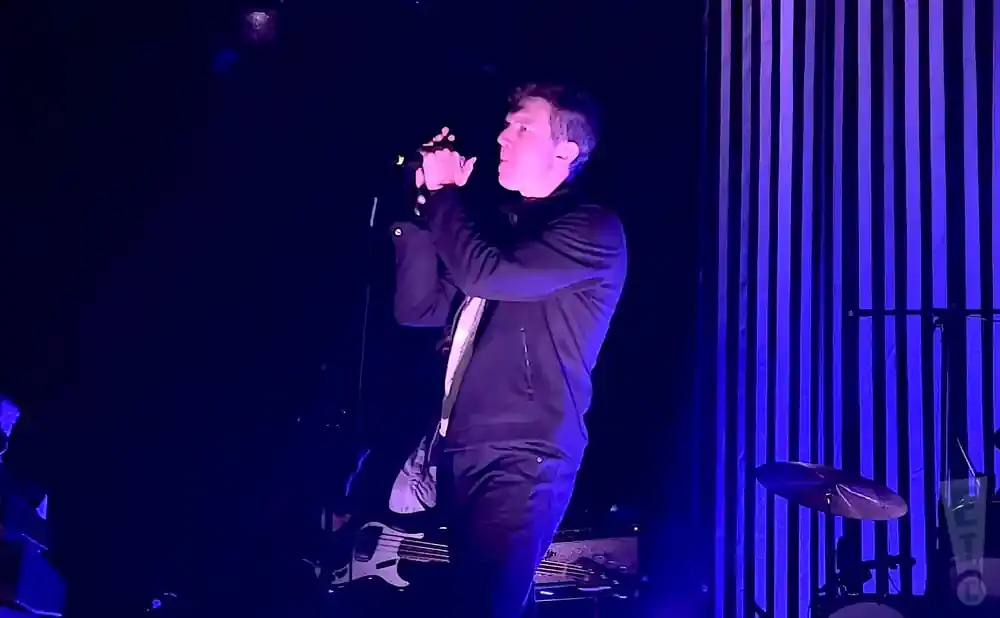 A man sings into a microphone on a dark stage with blue striped curtains behind him.