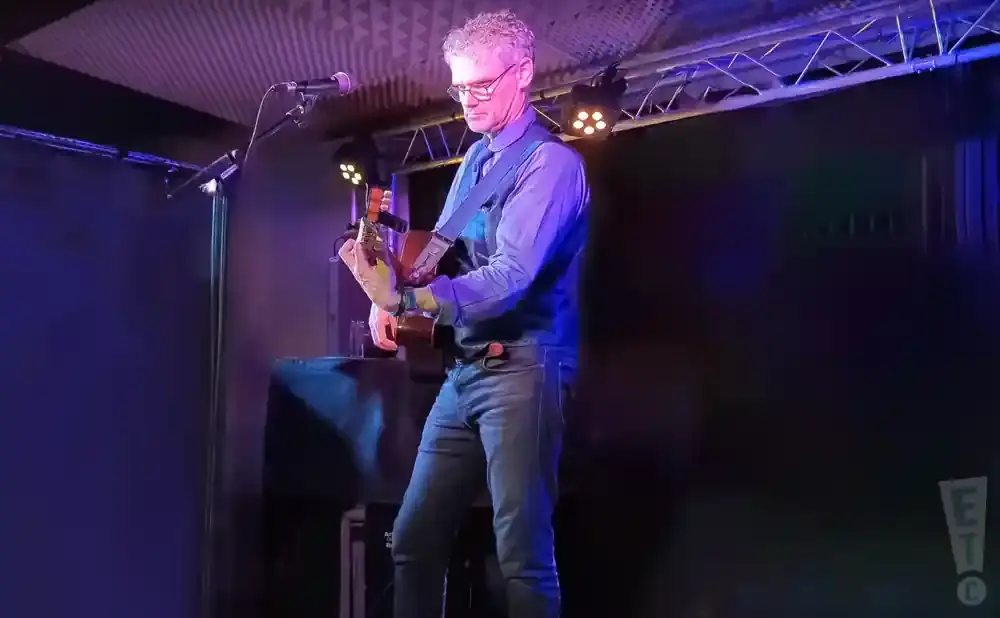 Man with gray hair and glasses playing an acoustic guitar on stage.