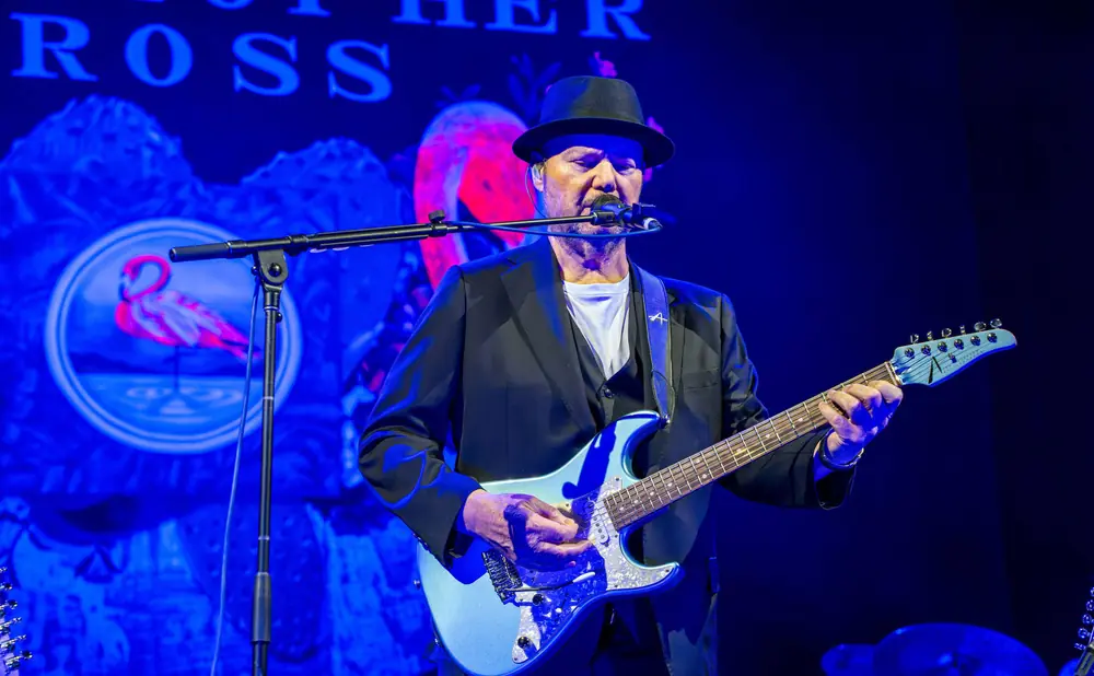 Musician in a hat and suit plays a blue guitar on stage with a flamingo logo backdrop.