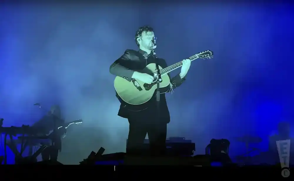 A man in a suit plays an acoustic guitar on a stage with blue lighting and smoke.
