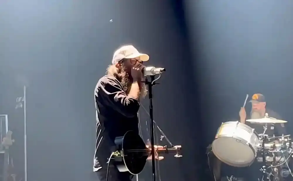 A man with a beard and glasses plays a harmonica into a microphone while a drummer plays in the background.