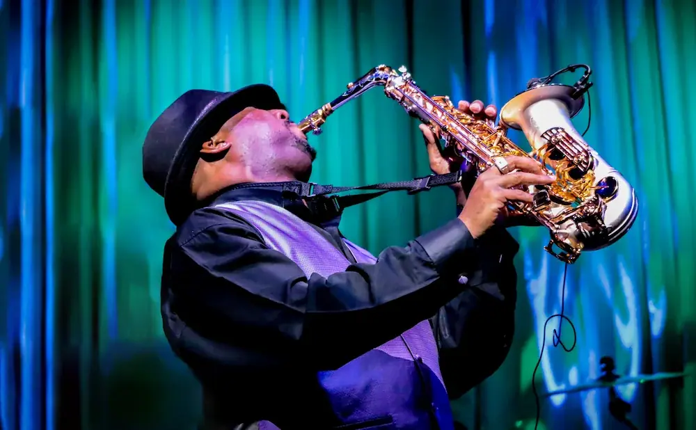 A man in a black hat and vest plays a saxophone on a stage with a blue and green backdrop.